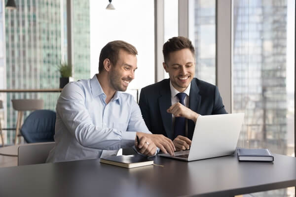 Two male workers, cheerful Latin and Caucasian multiethnic teammates cooperating on on-line project, sit at desk with laptop